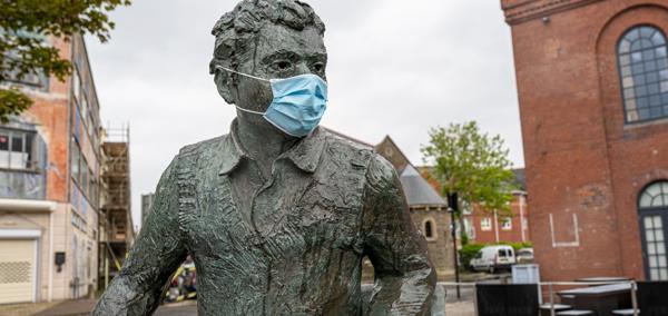 A bronze statue of Dylan Thomas seated outdoors in a paved courtyard, wearing a face mask, with brick buildings and trees in the background.