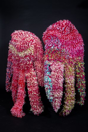 Two fluffy hand sculptures in front of a black background. The hands are primarily made out of pink yarn. Light blue, green, red and yellow pieces of yarn are also dotted throughout the objects.