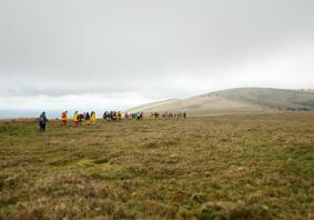 A photograph showing a grassy landscape with hills in the background. A group of schoolchildren wearing yellow raincoats and backpacks walk towards the hills. The sky above is cloudy and grey.