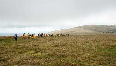 A photograph showing a grassy landscape with hills in the background. A group of schoolchildren wearing yellow raincoats and backpacks walk towards the hills. The sky above is cloudy and grey.