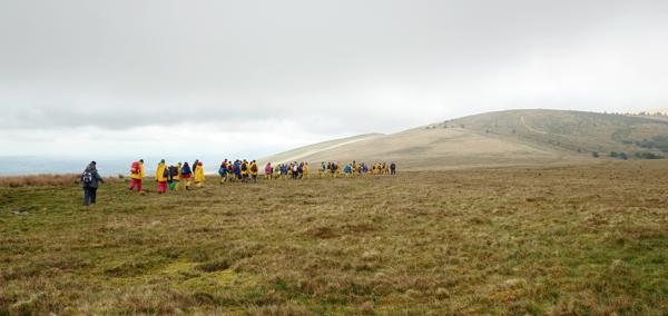 A photograph showing a grassy landscape with hills in the background. A group of schoolchildren wearing yellow raincoats and backpacks walk towards the hills. The sky above is cloudy and grey.