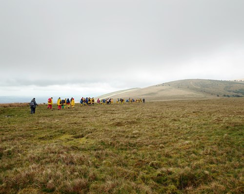 A photograph showing a grassy landscape with hills in the background. A group of schoolchildren wearing yellow raincoats and backpacks walk towards the hills. The sky above is cloudy and grey.