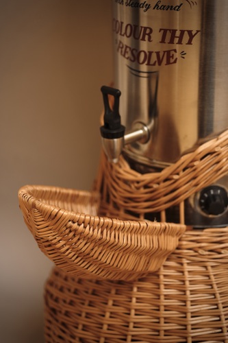 Wicker sculpture of a hybrid hot water dispenser encased in a basket structure, with a rounded bulbous base, open central column revealing internal components, and a small woven spout-like handle on the front.