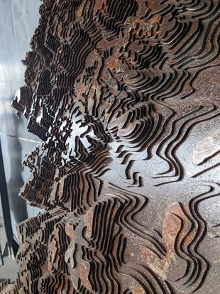 Close-up of the sculptural map, showing layered steel cut into flowing contour lines that create a textured, wave-like effect.