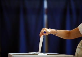 Female arm placing a ballot paper in a ballot box with a blue background.