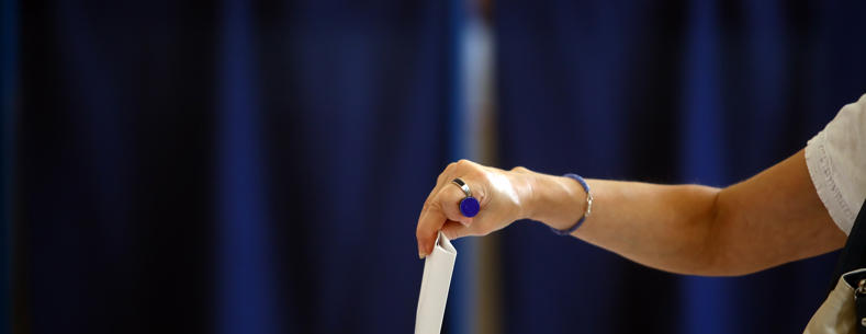 Female arm placing a ballot paper in a ballot box with a blue background.