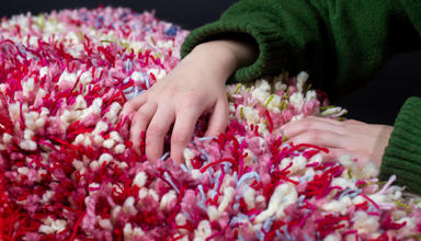A close up of a person holding a fluffy object. Different colours of yarn have been used including pink, green, red and blue.