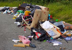 A pile of fly-tipped rubbish dumped on a road, including furniture, black bin bags, a sofa, cushions, toys, and household items scattered on the ground beside grass.