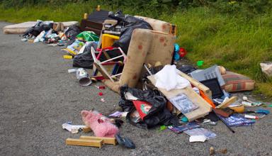 A pile of fly-tipped rubbish dumped on a road, including furniture, black bin bags, a sofa, cushions, toys, and household items scattered on the ground beside grass.