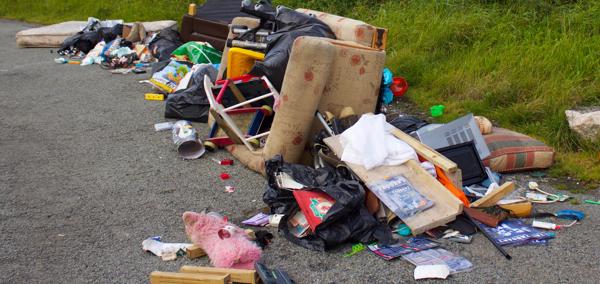 A pile of fly-tipped rubbish dumped on a road, including furniture, black bin bags, a sofa, cushions, toys, and household items scattered on the ground beside grass.