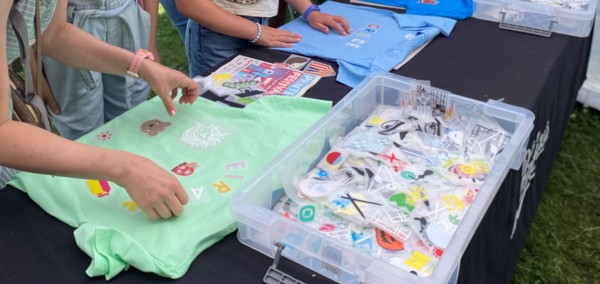A group of people gathered around a table covered with a black cloth, decorating coloured T-shirts using iron-on patches and letters.