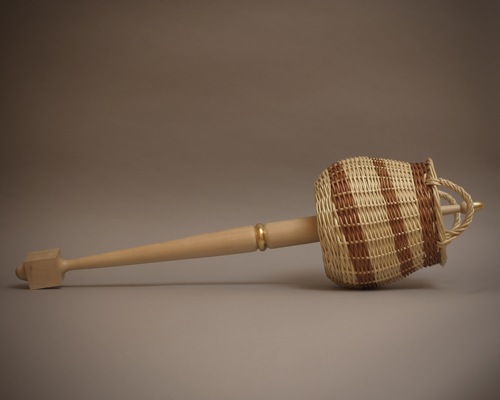 Wooden ceremonial mace lying on a plain neutral background, with a smooth light wood handle and a woven willow basket forming the head instead of a traditional royal crest, featuring a curved handle and subtle brown pattern in the weave.