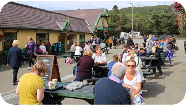 A photo showing people sitting outside Snowdon Mountain Railway Station. The area is busy with people sitting at several picnic tables and talking.