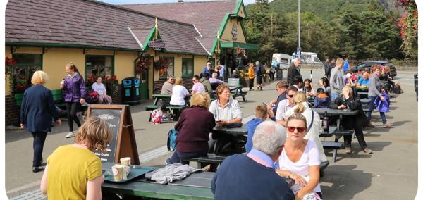 A photo showing people sitting outside Snowdon Mountain Railway Station. The area is busy with people sitting at several picnic tables and talking.
