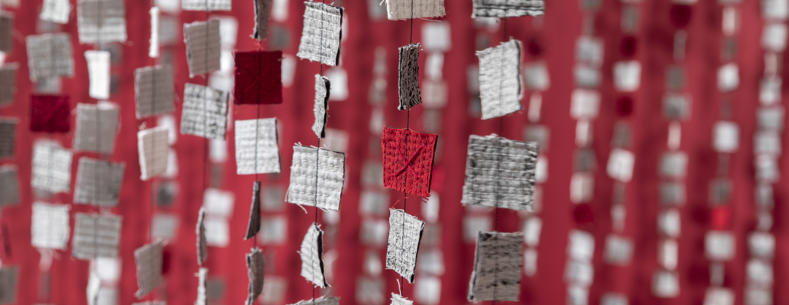 Rows of small square fabric pieces in white, grey, and red hang in vertical strands, creating a layered curtain effect against a red backdrop.