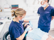 A dentist’s room with a patient wearing protective glasses lying back in the dentist chair. A dental worker wearing a mask, gloves and protective glasses is looking in the patient’s mouth. Another dental worker, also wearing a mask, looks on.
