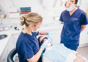 A dentist’s room with a patient wearing protective glasses lying back in the dentist chair. A dental worker wearing a mask, gloves and protective glasses is looking in the patient’s mouth. Another dental worker, also wearing a mask, looks on.