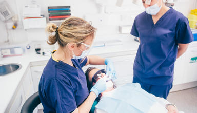A dentist’s room with a patient wearing protective glasses lying back in the dentist chair. A dental worker wearing a mask, gloves and protective glasses is looking in the patient’s mouth. Another dental worker, also wearing a mask, looks on.