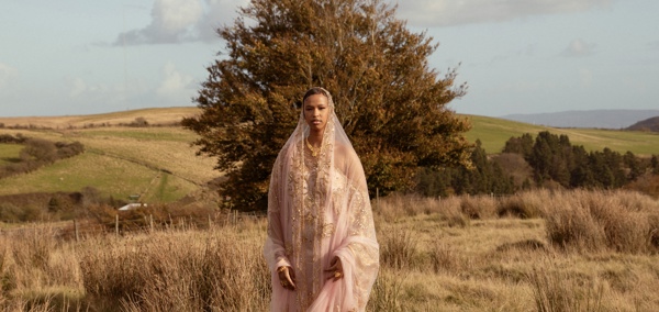 A young woman in a pink traditional Somali dress, standing in a grassy field with rolling hills and trees in the background.