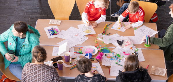 Children and adults gathered around tables covered in colourful art supplies, taking part in craft activities in a bright, spacious room.