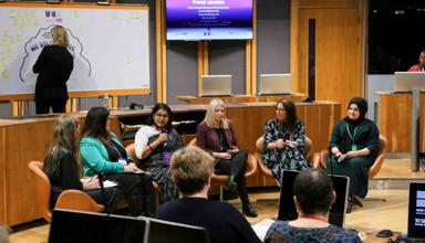 Six women sitting in the center of the Chamber in the Welsh Parliament, taking part in a conference and speaking to microphones.