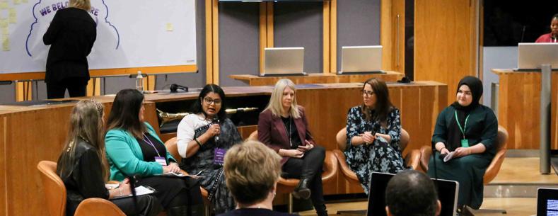 Six women sitting in the center of the Chamber in the Welsh Parliament, taking part in a conference and speaking to microphones.
