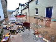 This is a picture of a street flooded by muddy water with floating rubbish and domestic items in the foreground. In the background are vehicles, most of which are submerged, one is  There is one floating car.