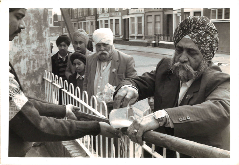 Person pours liquid from a metal jug into a container held by another person across a metal fence, with several people standing nearby on a residential street.