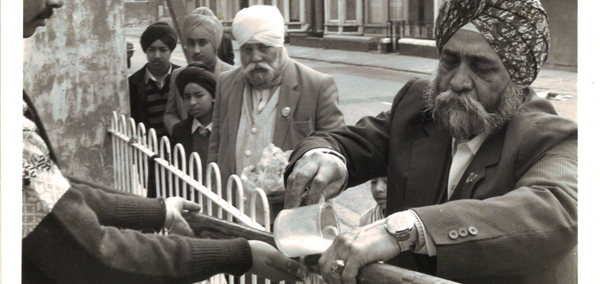 Person pours liquid from a metal jug into a container held by another person across a metal fence, with several people standing nearby on a residential street.