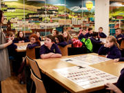A teacher speaks to a class of primary-age children. The children are listening, facing forwards. There is a large picture on the wall of mixed urban and rural landscapes.