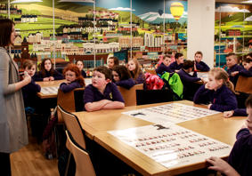 A teacher speaks to a class of primary-age children. The children are listening, facing forwards. There is a large picture on the wall of mixed urban and rural landscapes.