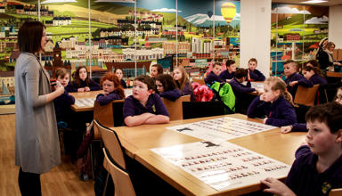 A teacher speaks to a class of primary-age children. The children are listening, facing forwards. There is a large picture on the wall of mixed urban and rural landscapes.