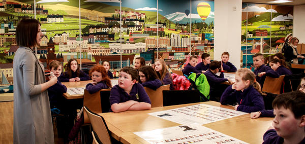 A teacher speaks to a class of primary-age children. The children are listening, facing forwards. There is a large picture on the wall of mixed urban and rural landscapes.